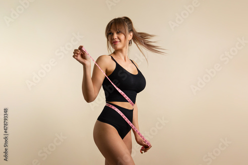 Slim healthy girl in black underwear with measuring tape at waist. Fit Woman poses in high waist panties and top. Perfect female body. Fitness, Diet or body care concept. Female shows her flat belly photo
