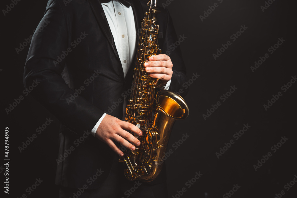 Naklejka premium Saxophonist Plays Jazz. Male Musician in a Formal Black Suit Holds a Tenor Saxophone on a Dark Background. Saxophone Close-up. Copy space High quality photo