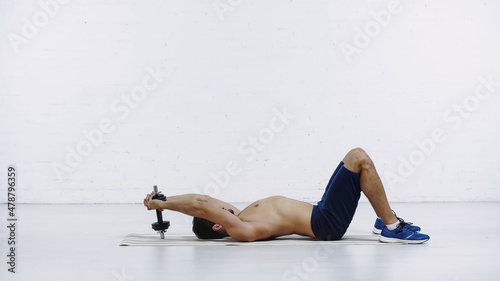 side view of shirtless sportsman in shorts training with heavy dumbbell while lying on fitness mat near white brick wall