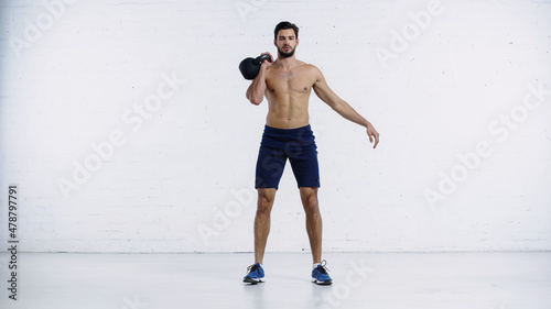 shirtless and sportive man exercising with heavy kettlebell near white brick wall