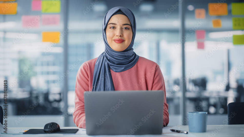Modern Office: Portrait of Young Muslim Businesswoman Wearing Hijab Works on Laptop, Does Data Analysis, Looks at Camera and Smiles. Empowered Digital Entrepreneur Works on e-Commerce Startup Project - obrazy, fototapety, plakaty 