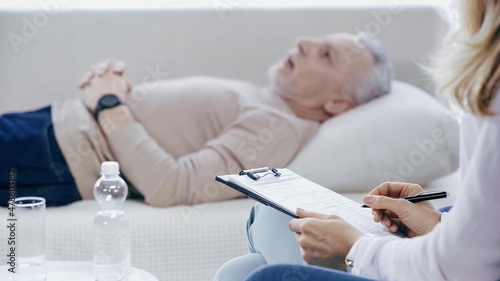 psychologist holding clipboard near middle aged man lying on sofa during therapy