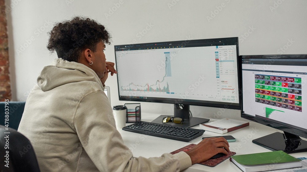 Concentrated young male trader looking at computer screen, studying charts while trading from home