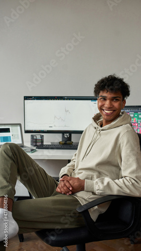 Cheerful young guy, mixed race trader work from home using laptop and computer, sitting in modern living room office