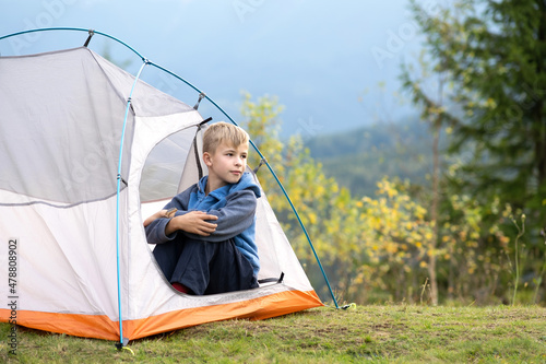 Hiker child boy resting sitting in a camping tent at mountains campsite enjoying view of summer nature