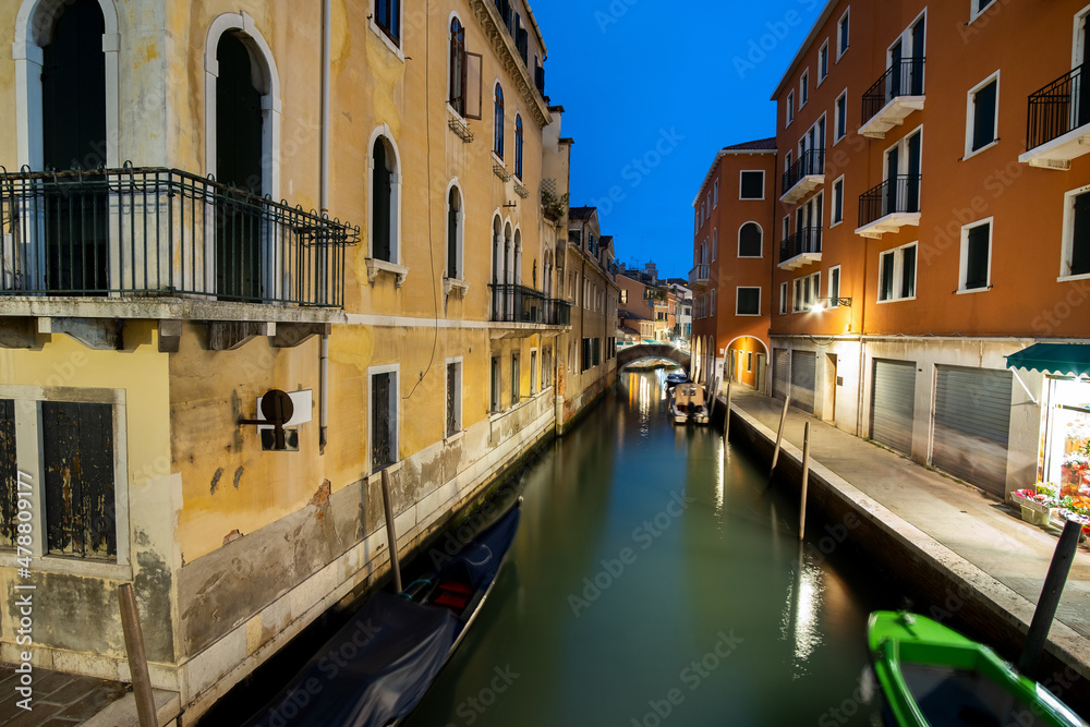 Night scene of illuminated old buildings, floating boats and reflections in canal water in Venice, Italy.