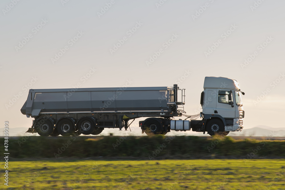 Fototapeta premium Semi-truck with tipping cargo trailer transporting sand from quarry driving on highway hauling goods in evening. Delivery transportation and logistics concept