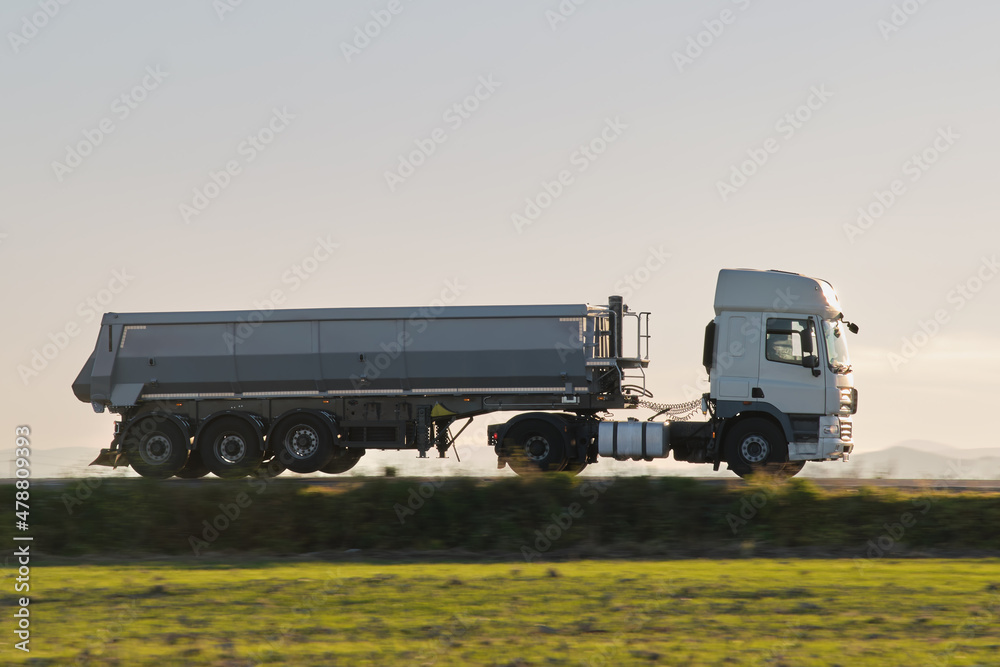 Semi-truck with tipping cargo trailer transporting sand from quarry driving on highway hauling goods in evening. Delivery transportation and logistics concept