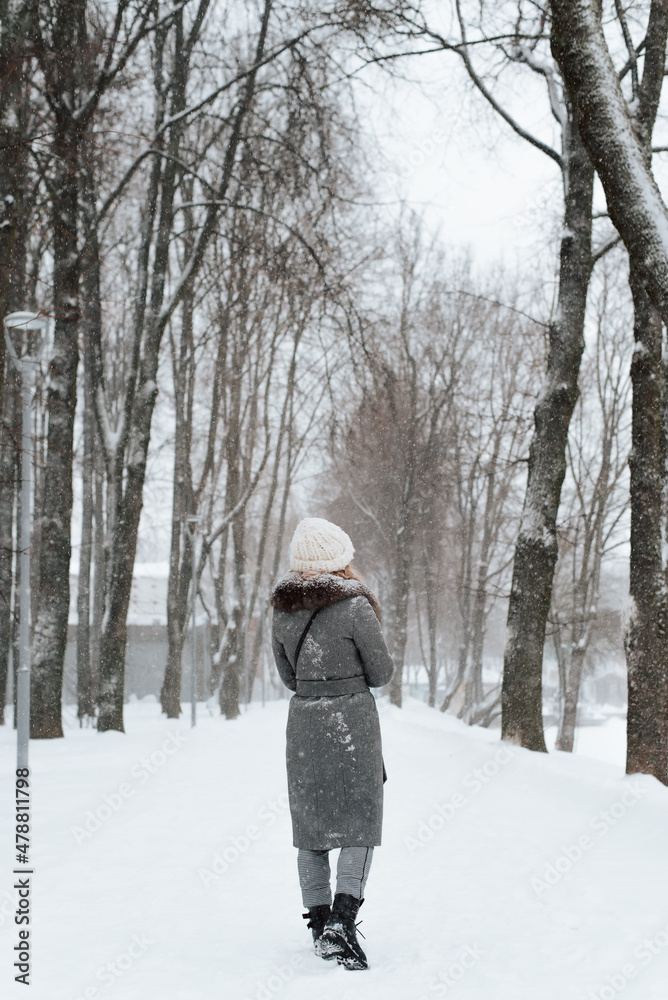 Back view of young woman in coat and knitted white hat enjoying walk in winter park, outdoors. Rear view of lonely girl walking along snowy alley. Vertical image