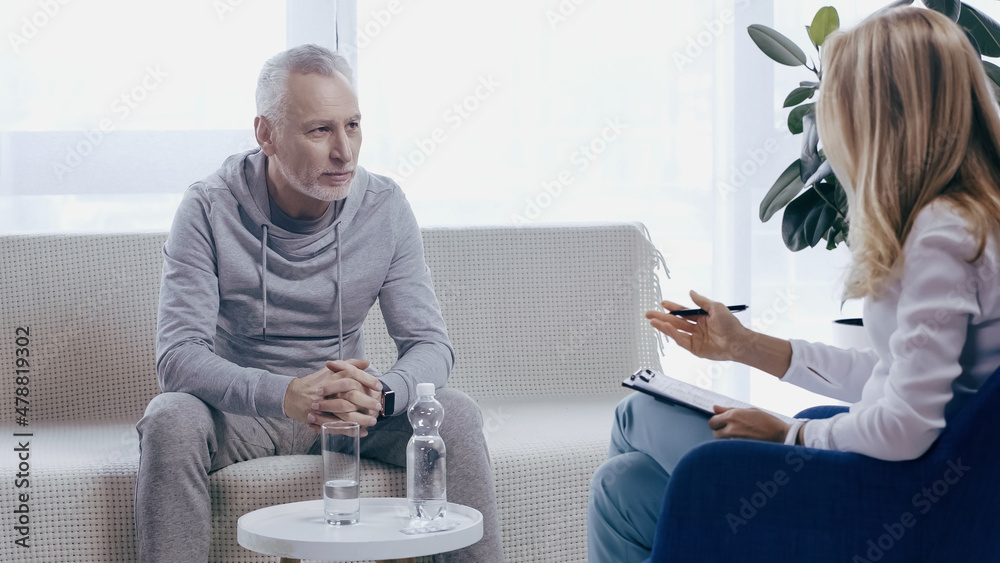 Obraz premium middle aged man in sportswear with clenched hands sitting on sofa near psychotherapist in consulting room