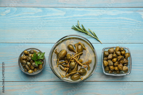 Tasty capers, rosemary and parsley on light blue wooden table, flat lay