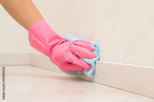 Young adult woman hand in pink rubber protective glove using blue dry rag and wiping light wooden baseboard surface in room at home. Closeup. photo