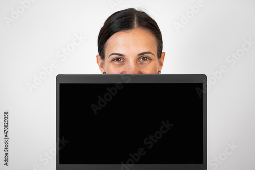 Brunette holding lap top in front of her face. Blank black screen in focus. Photo taken on grey background.