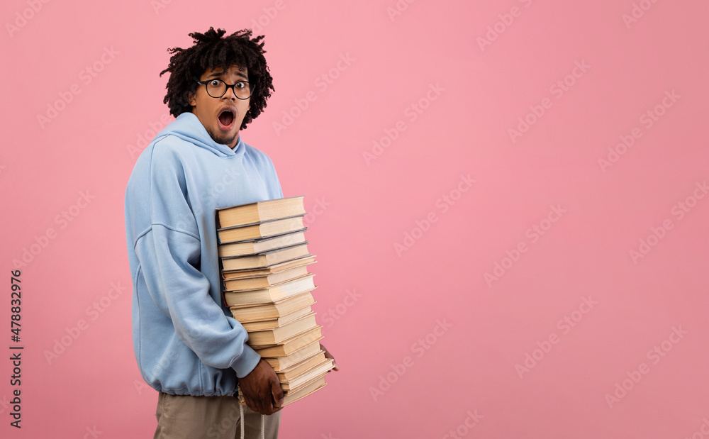 Nerdy black teenage student feeling shocked over pile of homework, holding big stack of books on pink background Nerdy black teenage student feeling shocked over pile of homework, holding big stack of books on pink background
