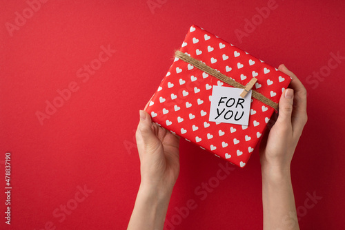 First person top view photo of valentine's day decor woman's hands holding giftbox in red wrapping paper with heart pattern twine and pinned note on isolated red background with blank space © ActionGP