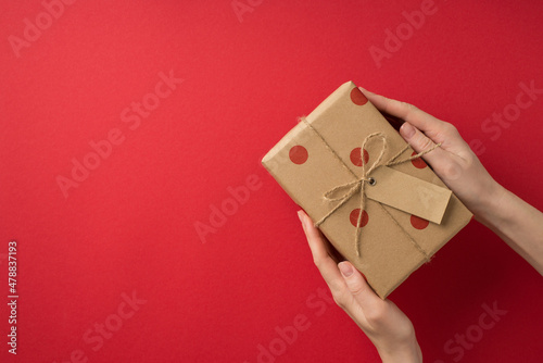 First person top view photo of saint valentine's day decorations young female hands taking kraft paper gift box with polka dot pattern and twine bow on isolated red background with blank space