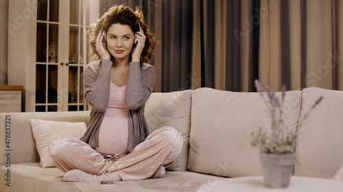 Young pregnant woman using headphones near cellphone on couch