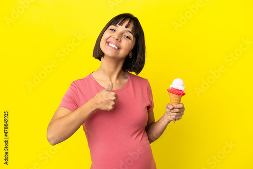 Pregnant woman holding a cornet ice cream isolated on yellow background giving a thumbs up gesture © luismolinero