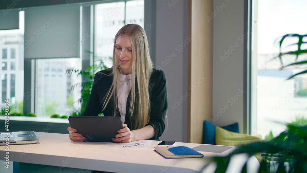 Elegant woman in suit sitting at table with laptop and surfing tablet in modern light office with big windows Elegant woman in suit sitting at table with laptop and surfing tablet in modern light office with big windows
