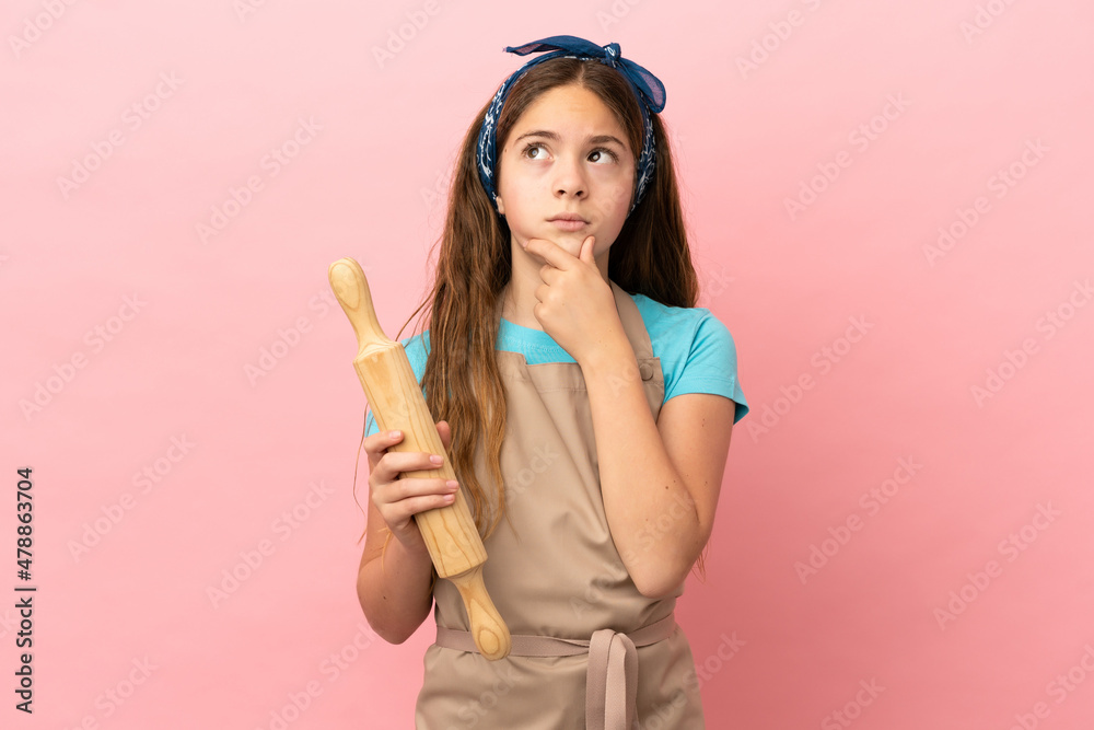 Little caucasian girl holding a rolling pin isolated on pink background having doubts Little caucasian girl holding a rolling pin isolated on pink background having doubts
