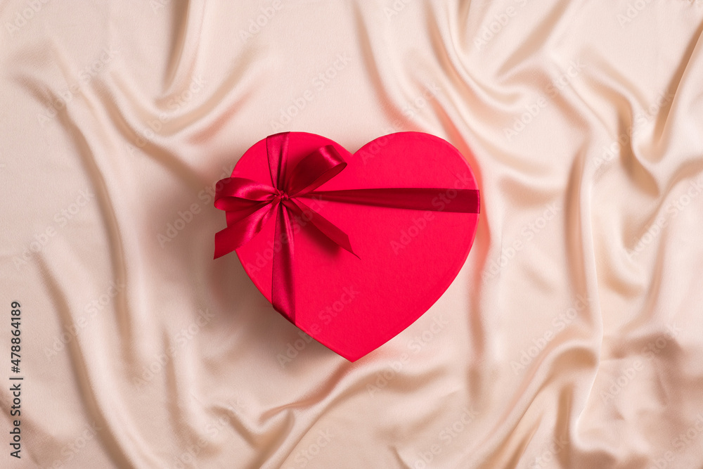 Red heart-shaped box wrapped ribbon now on silk background. Happy Valentines Day concept. Flat lay, top view. Red heart-shaped box wrapped ribbon now on silk background. Happy Valentines Day concept. Flat lay, top view.