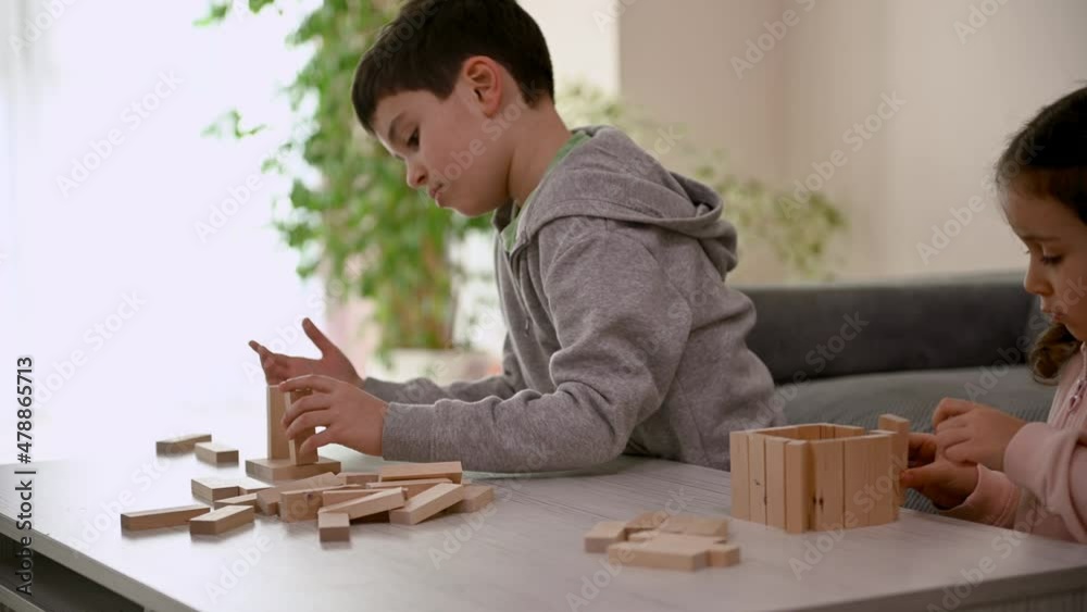 Cute children concentrated on building with wooden blocks. Little girl rejoices when she finishes construction, talks to her brother who pays no attention to her, focusing on building a tall tower Cute children concentrated on building with wooden blocks. Little girl rejoices when she finishes construction, talks to her brother who pays no attention to her, focusing on building a tall tower