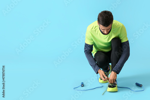 Sporty young man with jumping rope tying shoelaces on color background