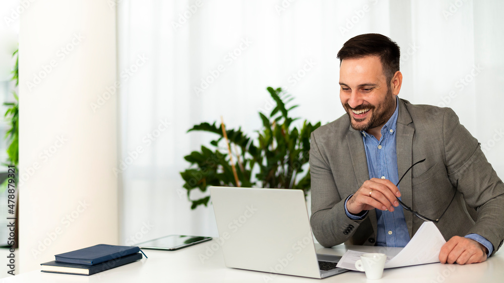 Smiling attractive young manager with a stylish short beard, sitting at his office desk at a online meeting with his team where good business results are announced