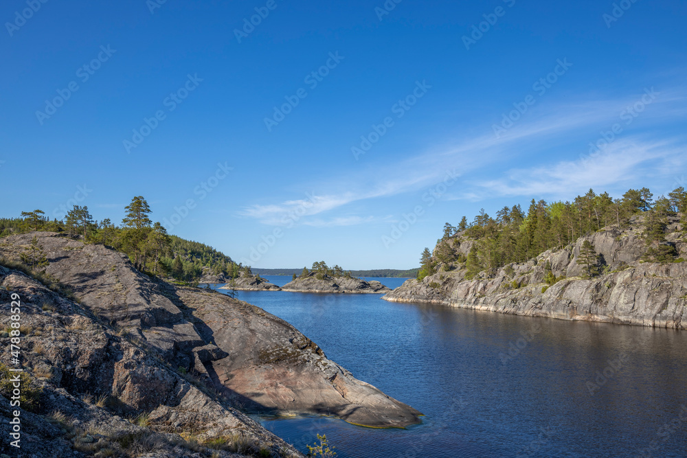 Fototapeta premium Landscape with a forest on stones over the lake. Sunny day at the lake. Reflection of the sky in the water. Pines on stones. The nature of the north.