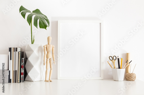 Poster frame mock up and leaf in geometric vase, clock and drawing tools on white table. Home office decor.