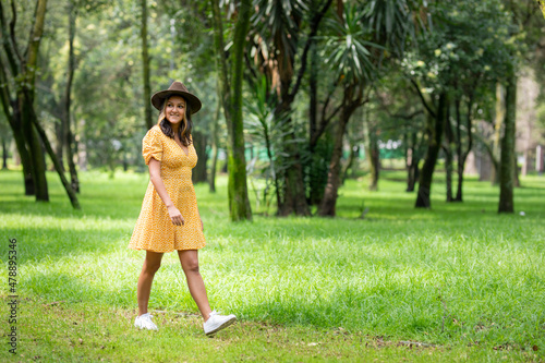 Young happy Hispanic woman walking through the park wearing a yellow dress and hat