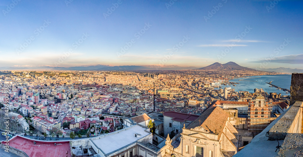 Wide angle view of City of Naples, mount Vesuvius and Gulf of Naples from the top of San Elmo Castle in the evening. Blue sky and sunset shadows on the old town. Ships in the port