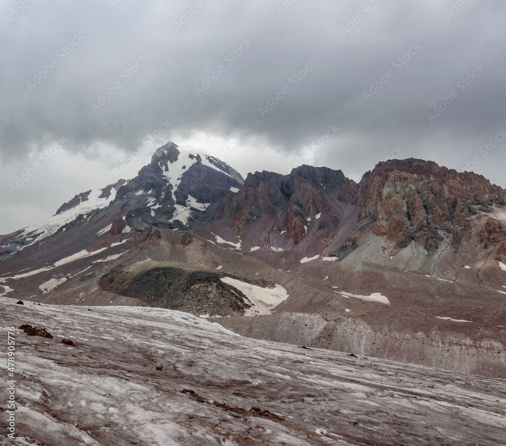Gergeti glacier and mount Kazbek in Georgia, grey thunder sky with clouds, grey and brown stones and rocks, and white stone on the slopes
