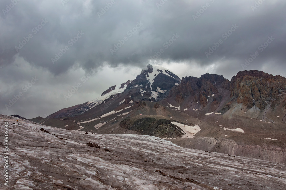 Gergeti glacier and mount Kazbek in Georgia, grey sky with clouds, grey and brown stones and rocks, and white stone on the slopes