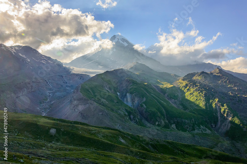 Green hills grey rocks and Mount Kazbek summit in Georgia under blue sky with clouds