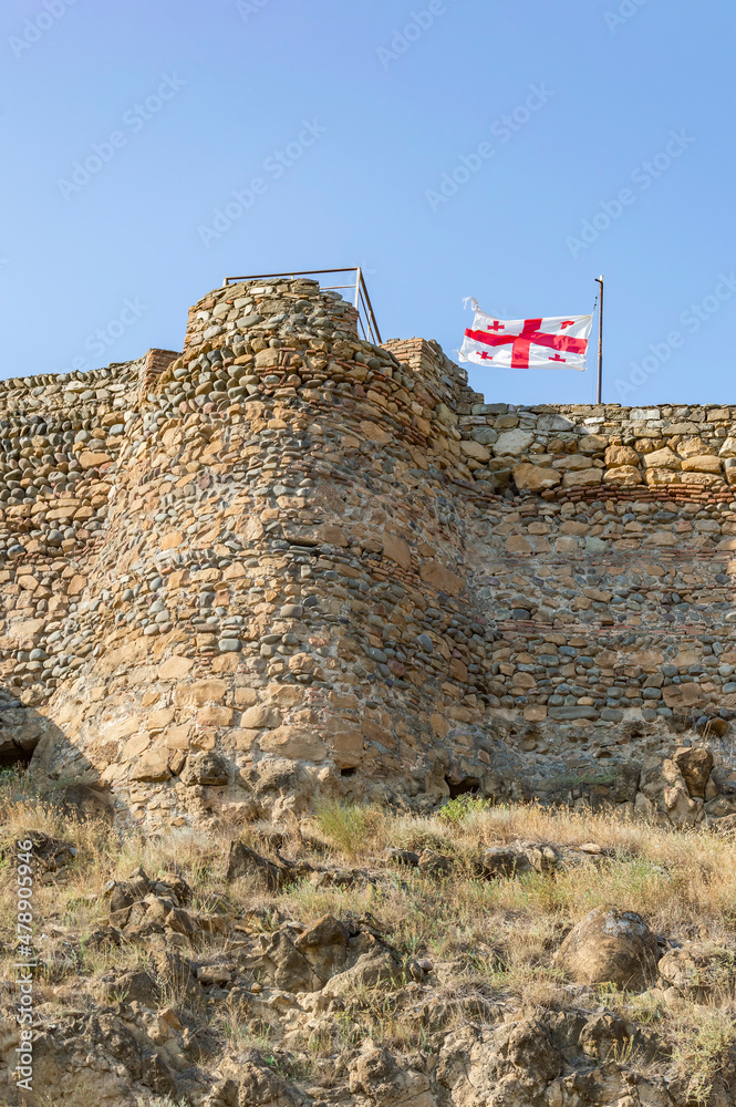 Obraz premium Georgian white and red flag with cross flying in the wind atop the medieval fortress in the town of Gori, Georgia. Ancient yellow, grey and brown stone walls and tower, dry yellow grass, blue sky
