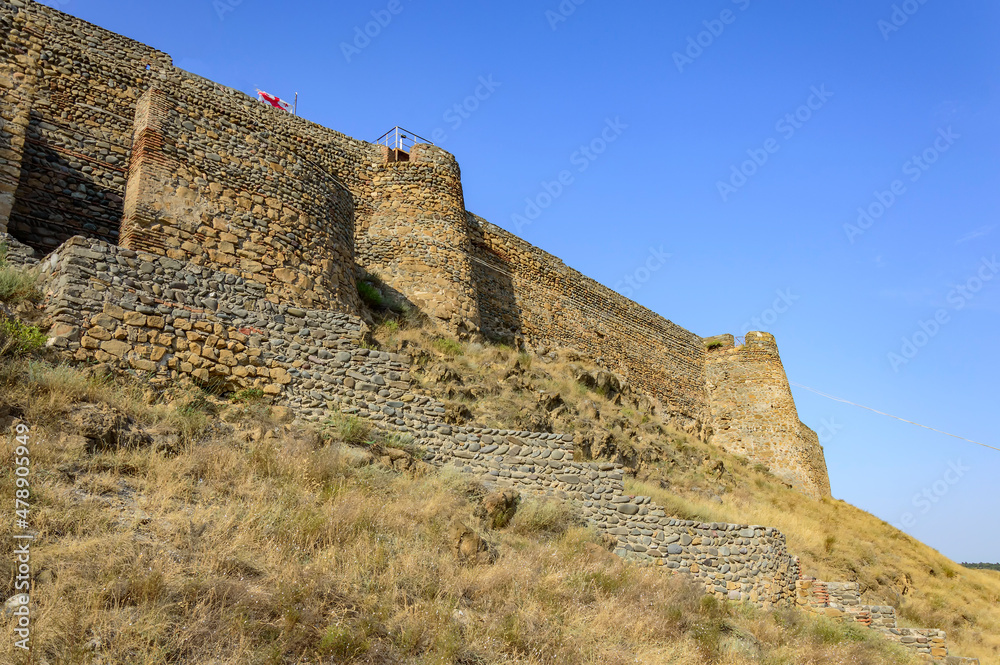 Medieval fortress in the town of Gori, Georgia. Ancient yellow, grey and brown stone walls and towers, dry yellow grass, blue sky