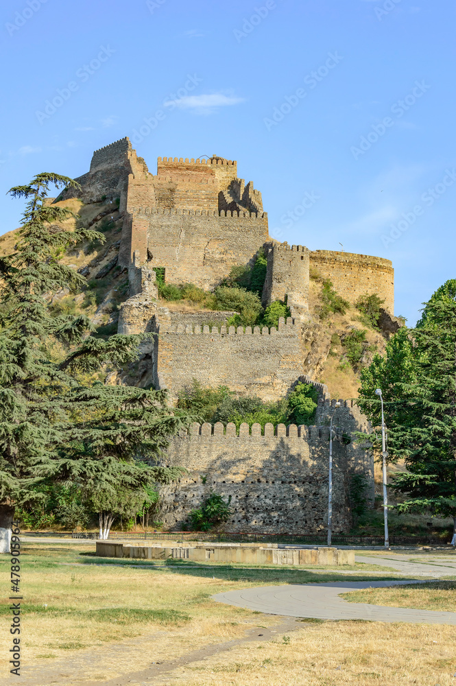 Medieval fortress in the town of Gori, Georgia. Ancient yellow, grey and brown stone walls and towers, green trees on the rocky slopes, blue sky with clouds, asphalt road