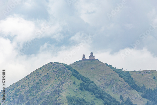 Caucasus mountains and Gergeti medieval church near Mount Kazbek in Georgia. Brown and green mountain slopes, sky with clouds, ancient church and bell tower walls photo