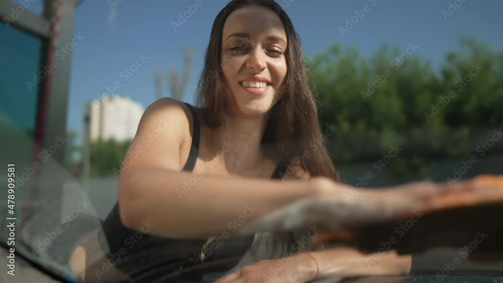 Smiling seductive slim beautiful woman cleaning windshield glass in slow motion. Portrait of brunette female employee washing automobile in slow motion at car wash service outdoors Smiling seductive slim beautiful woman cleaning windshield glass in slow motion. Portrait of brunette female employee washing automobile in slow motion at car wash service outdoors