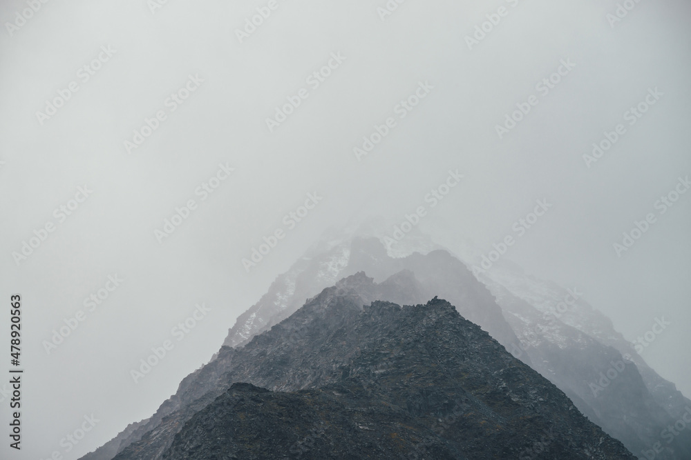 Dark atmospheric landscape with high black rocky mountain top with snow in cloudy sky. Dramatic mountain landscape with snow peaked top in haze in overcast weather. Gloomy view to awesome rocks in fog