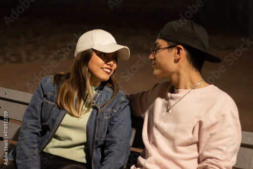 young couple in the park sitting on a bench in the evening looking lovingly at each other, they have an urban clothing style © Carlos
