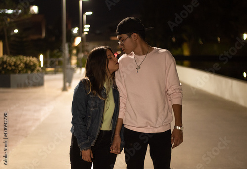 young latin couple walking in the street at night holding hands, looking at each other with love, illuminated by the city lights and wearing urban style clothes.