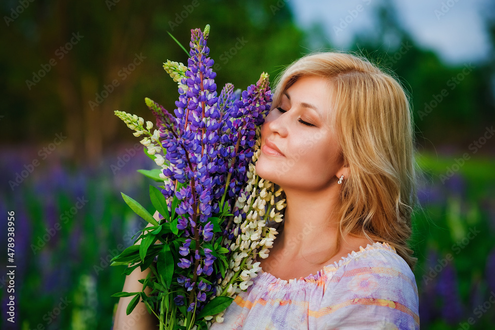 Portrait of a beautiful plus-size blonde with a bouquet of lilac-purple lupine flowers in her hands. A stout woman stands on a blooming meadow. Portrait of a beautiful plus-size blonde with a bouquet of lilac-purple lupine flowers in her hands. A stout woman stands on a blooming meadow.