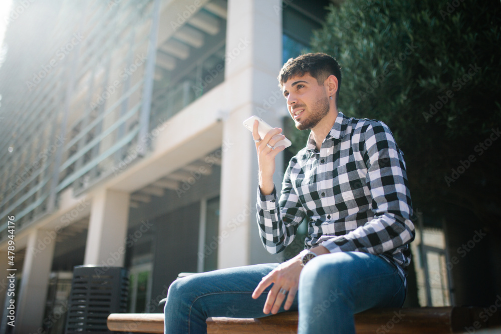 Fototapeta premium young, handsome guy sitting on a bench and sending an audio note with his phone