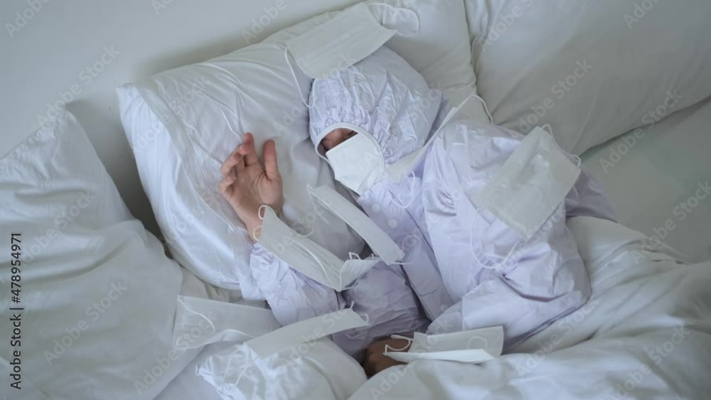 Paranoid person in protective uniform sleeping in bed, ironically covered with lots of face masks, covid-19 quarantine Paranoid person in protective uniform sleeping in bed, ironically covered with lots of face masks, covid-19 quarantine