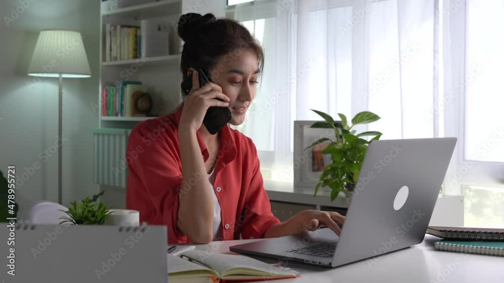 Beautiful young Asian woman work at home with laptop and talking on smartphone and smiling. Female working on computer and using cell mobile phone while sitting at home Beautiful young Asian woman work at home with laptop and talking on smartphone and smiling. Female working on computer and using cell mobile phone while sitting at home