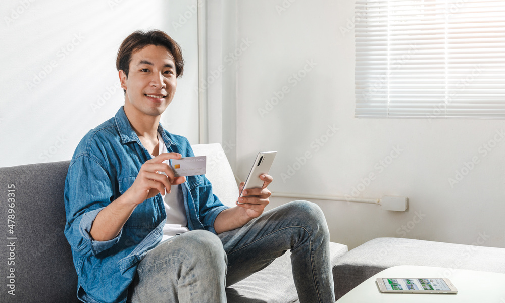 Happy millennial young Asian man sitting on couch at home or co-working space using a credit card and cellphone for online shopping. Technology money wallet and transferring money app payment concept Happy millennial young Asian man sitting on couch at home or co-working space using a credit card and cellphone for online shopping. Technology money wallet and transferring money app payment concept