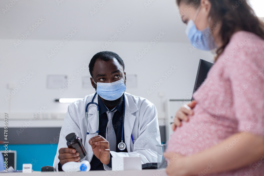 Practitioner doing consultation with pregnant patient, wearing face masks for protection. Woman expecting child and receiving medical advice about pregnancy from doctor during pandemic Practitioner doing consultation with pregnant patient, wearing face masks for protection. Woman expecting child and receiving medical advice about pregnancy from doctor during pandemic