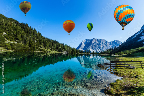 Colorful hot air balloons flying over the lake surrounded by mountains. large multi-colored balloons slowly rising against blue sky. Travel, adventure, festival.
